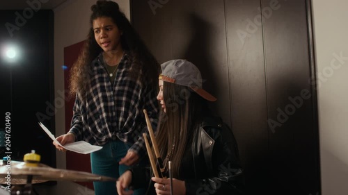 Caucasian female teenager practicing drums while African American female teenager holding sheet music explaining rhythm during rehearsal. Clip is perfect for music school advertising