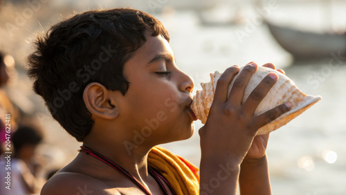 Young indian boy participates in community ceremony by blowing conch shell near water, symbolizing tradition and cultural spirit