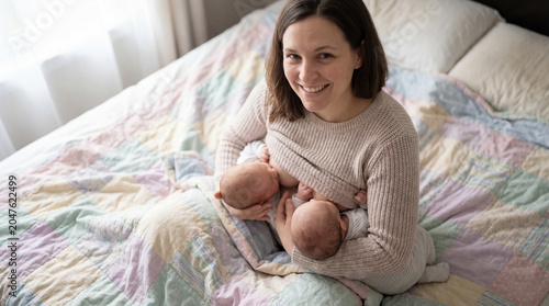 Smiling mother nurses twin infants on bed showing postpartum bonding tandem breastfeeding newborn care family love maternal wellness supportive home life early parenting gentle newborn routine