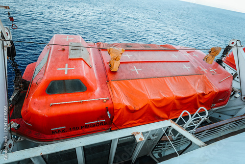 Foto High angle view of an orange enclosed emergency lifeboat marked for maximum 150 persons, mounted on a passenger vessel deck with the blue sea in the background
