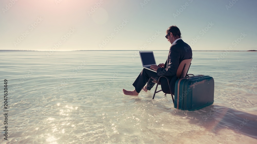 Fototapeta premium Businessman in suit working on laptop while sitting on chair in shallow ocean water next to a suitcase, representing work life balance