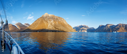 Panorama of the icebergs, hanging glaciers and rugged mountains of Kjerulf Fjord in Northeast Greenland National Park. Seen from a ship, with deep blue water and a summer sky background.