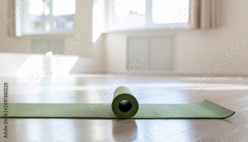 Green yoga mat rolled on light wooden floor in bright sunlit studio with blurred window background