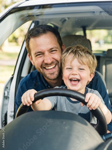 Close up of laughing father and little son looking through car windshield holding steering wheel together