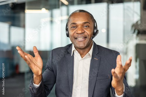 Mature man wearing a headset  smiling and gesturing while communicating during a virtual business meeting or providing customer support from a modern office environment