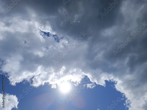 Bright sun emerging from behind dark grey storm clouds. Dramatic weather background with blue sky