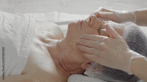 Cosmetologist's hands rubbing rejuvenating oil on the face and body of an elderly woman