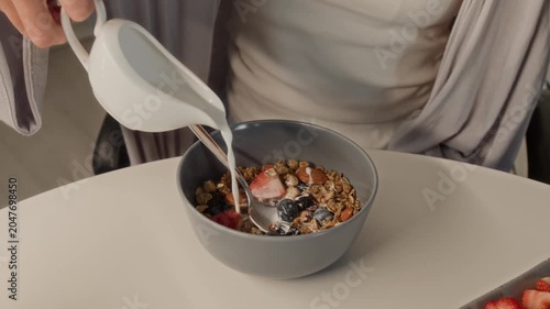 Close up on hands of young adult woman pouring milk from pitcher into bowl of granola with fresh berries and stirring breakfast. Clip perfect for healthy lifestyle and organic food commercials
