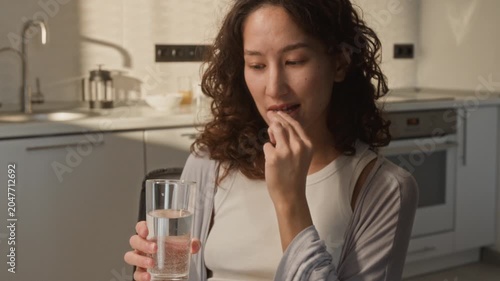 Tilt up of Asian young adult woman taking vitamin capsule from pill organizer and drinking water during breakfast. Footage perfect for wellness, healthcare, and daily supplement commercial campaigns