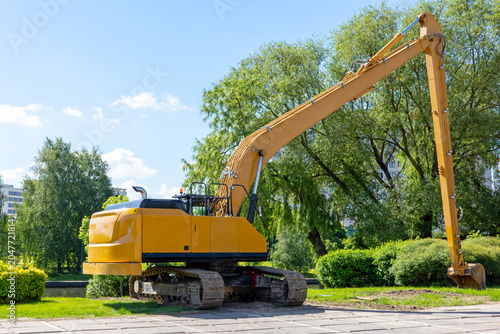 Slika na platnu A large yellow construction excavator with an extended long-reach boom stands stationary on a paved surface, ready for heavy-duty earthmoving and demolition work outdoors