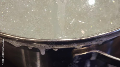 Boiling liquid with foam on surface in a metal pot. Process of cooking food in domestic kitchen. Close up view of water bubbling in stainless steel cookware. Culinary preparation concept.