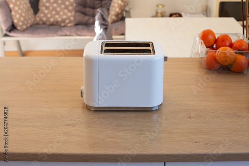 Minimalist white toaster standing on a wooden kitchen counter in a modern home kitchen. Breakfast appliance and household equipment concept