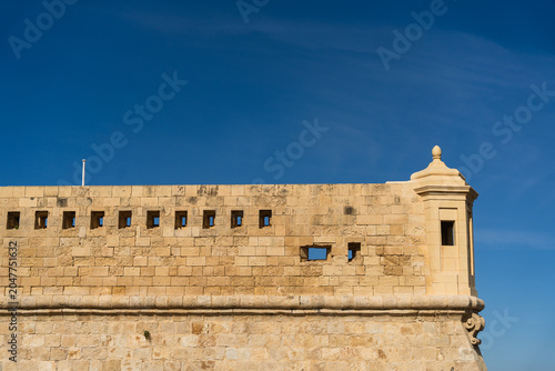 View of the historic limestone fortification walls and a traditional watchtower under a clear blue sky in Valletta, Port Region, Malta.