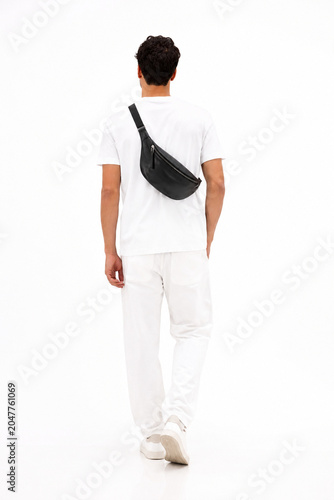 Rear view of a young man walking away on a white background. She is wearing a white t-shirt, white pants, and white sneakers, carrying a black leather crossbody bag. Minimalist studio shot ideal for