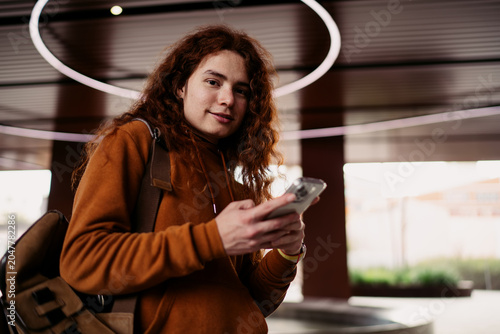 Smiling man with curly hair looking at camera holding smartphone, male freelancer using mobile banking app outdoors, authentic representation of young adult with cellphone, 5G data, Caucasian model.