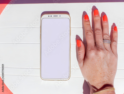 Minimalist flat lay of a smartphone, a single hand reaching toward it from one corner--clean composition with color-pop nails or bracelet, white tabletop background