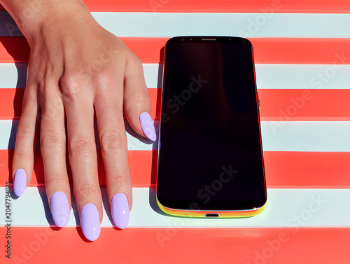 Minimalist flat lay of a smartphone, a single hand reaching toward it from one corner--clean composition with color-pop nails or bracelet, white tabletop background
