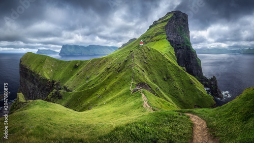 View of Kallur Lighthouse on a steep green cliff with a narrow dirt path and dramatic clouds over the ocean Klaksvik, Northern Isles, Faroe Islands.