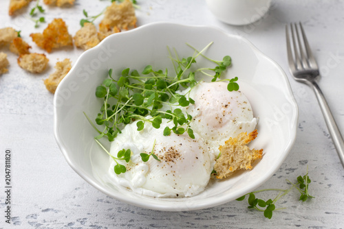 Poached eggs with microgreens and bread croutons in white bowl on light rustic background