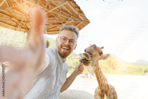 Fényképezés Young caucasian male feeding giraffe in zoo on sunny day in amusement park Nha t