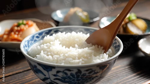 Steaming hot white rice being scooped from a bowl with a wooden spoon, served with side dishes