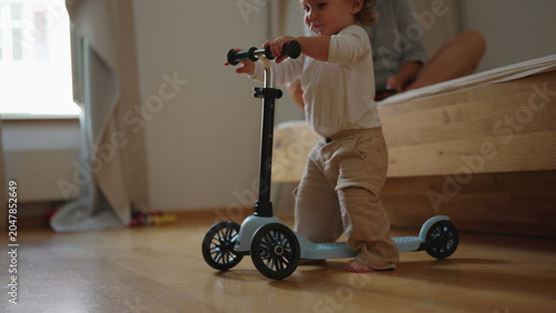 Foto Toddler learning to ride small scooter indoors while repeatedly falling and gett