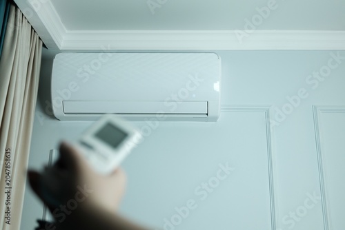 Photos Hand using a remote control to operate a modern wall-mounted air conditioner in a bright home interior