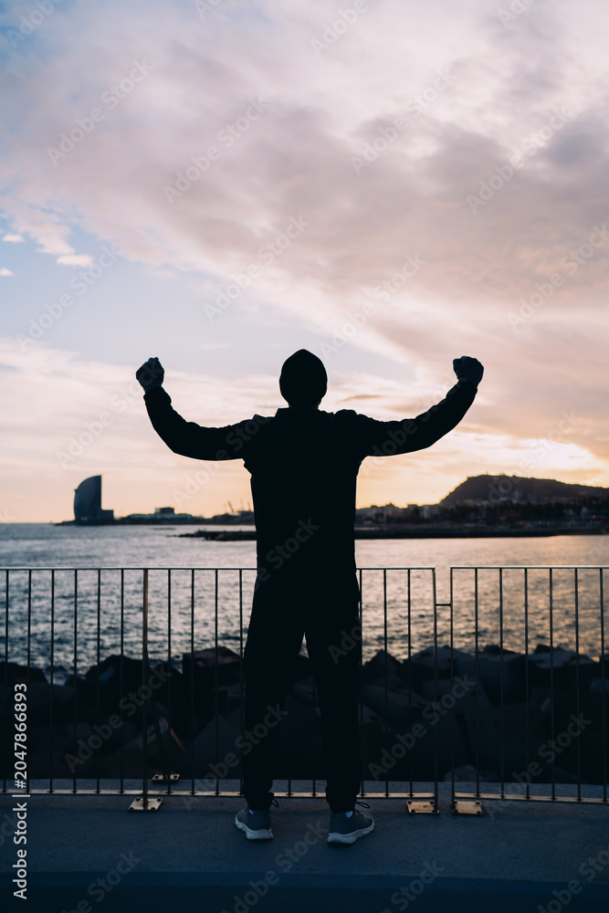 Fototapeta premium Athlete standing on modern bridge at sunset, man in black sportswear celebrating success with raised arms, triumph and achievement concept, urban wellness and fitness motivation, harbor view.