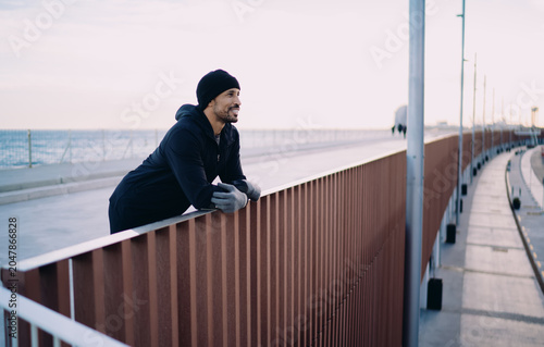 Obraz na plátně Content man leaning on railing of modern sea port walkway, athlete resting during morning workout, urban wellness and mindfulness, healthy active lifestyle, peaceful city view at harbor