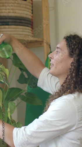 Vertical video: Lifting green bottle, person misting plant to care at home in white top, copy space