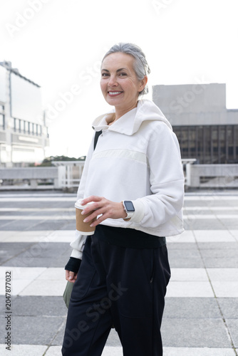 Holding paper cup, senior wearing white hoodie carrying olive bag on rooftop plaza with smartwatch