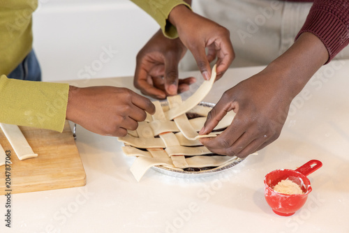 African American friends weaving lattice over fruit pie in aluminum tin, red measuring cup visible