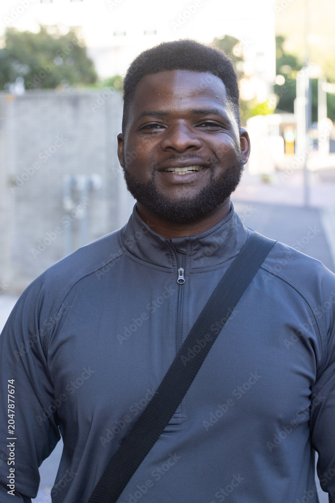 Naklejka premium African man standing on urban sidewalk smiling at camera wearing dark-gray pullover and black strap