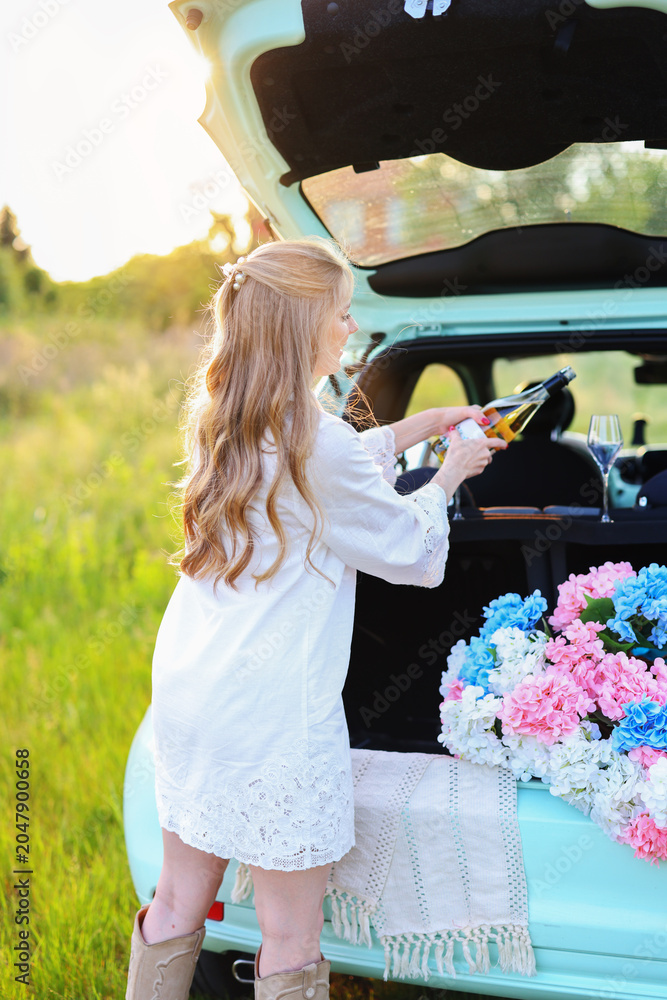 Fototapeta premium Elegant blonde woman serving wine from the open trunk of a compact car decorated with hydrangeas during a warm sunset in nature