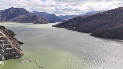 Aerial tracking shot pulling back over the vast, pale-green waters of the Moglicë reservoir, framing the surrounding steep valleys, rocky slopes, and distant mountain ridges in Albania