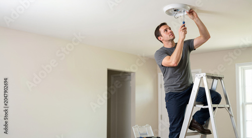 Man standing on stepladder installing ceiling light fixture with screwdriver. Electrician repairing electrical wiring in empty room. Home renovation concept