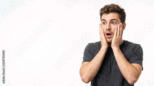 A young man with a startled and wide-eyed expression, holding his hands to his face in genuine shock or intense surprise against a clean white background