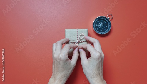Hands tying a small gift box with twine on red surface, beside a compass for navigation