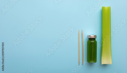 Minimalist composition featuring celery, green juice vial, and toothpicks against light blue backdrop