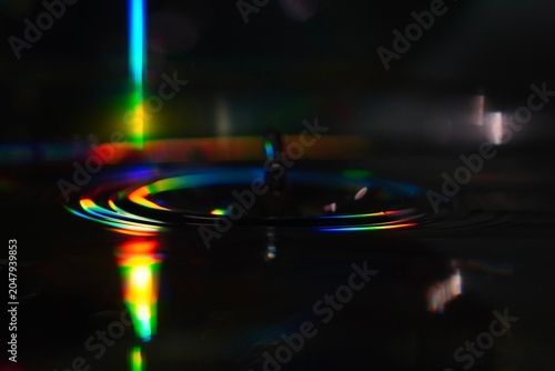 An aesthetic, close-up conceptual shot of water ripples. Vibrant rainbow light leaks, refraction, and spectral colors create a futuristic, neon, and meditative atmosphere against a deep black backdrop