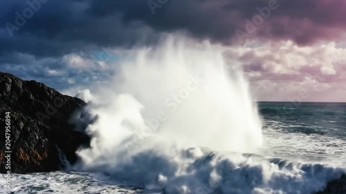Powerful ocean waves crashing against rocky cliffs under a dramatic cloudy sky on a stormy day, spraying water and foam.
