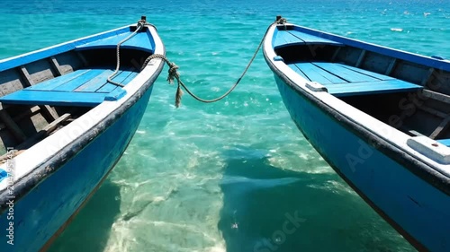 Two connected blue boats gently float on clear turquoise water in the bright sunlight.