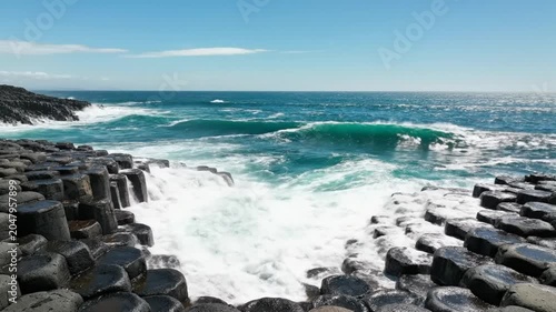Waves crashing against hexagonal basalt columns with turquoise water under a blue summer sky