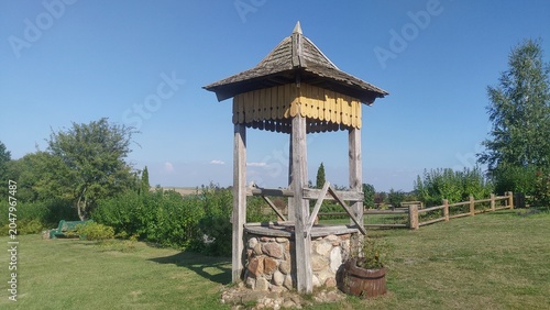 Tableau sur toile There is a stone well with a wooden superstructure and a shingled roof on a grassy lawn