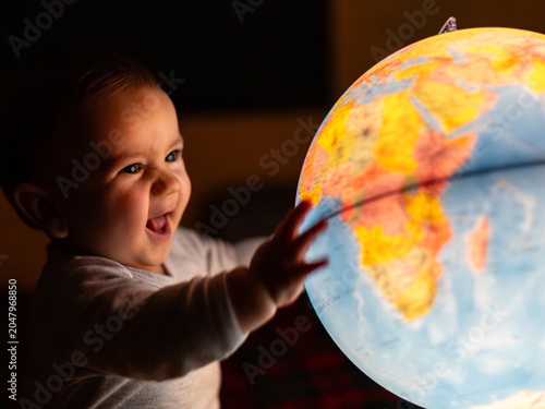 Happy baby smiling and reaching hand to touch illuminated world globe lamp in dark room discovering earth geography concept for childhood education curiosity.