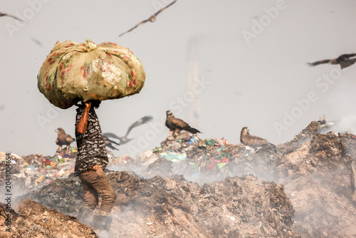 Aminbazar, Bangladesh - 21 February 2026: View of a waste picker carrying a large sack on his head amidst mounds of garbage and scavenging birds at the Aminbazar landfill.