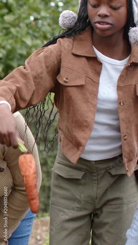 Vertical video: Seeing greens, two girls in knit hats pulling carrot in garden bed, sharing harvest