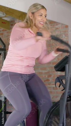 Vertical video: Gripping handles woman pedaling airbike at home for fitness raising arm, pink top