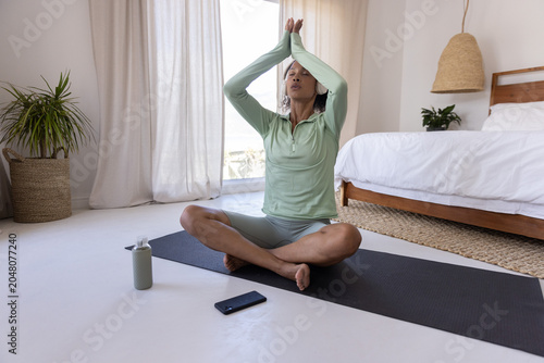 Black yoga mat lying on hardwood floor near sheer window curtains, with water bottle and smartphone