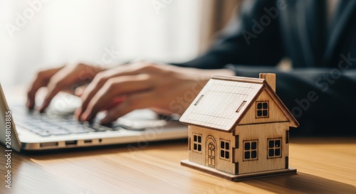 Hand holding miniature wooden house model over laptop on wooden desk with natural light